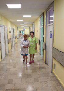 korytarz oddziału szpitala koloru żółtego z szarą podłogą uśmiechają się dwie osoby ubrane w odzież ochronną koloru zielonego i białego