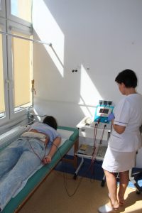 terapeuta ubrany w białe ubranie medyczne ochronne wykonuję zabieg rehabilitacyjny pacjętowi na łóżku medycznym koloru zielonego aparatem koloru białego