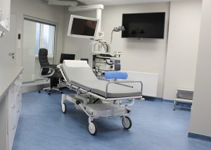 pokój badań endoskopowych w którym znajduje się łóżko medyczne koloru szarego, stołek obrotowy koloru niebieskiego, na scianie ekran koloru czarnego wraz z aparaturą do badań endoskopowych