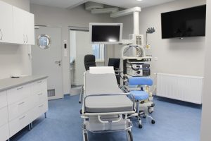 pokój badań endoskopowych w którym znajduje się łóżko medyczne koloru szarego, stołek obrotowy koloru niebieskiego, na scianie ekran koloru czarnego wraz z aparaturą do badań endoskopowych