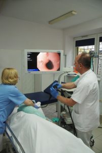 lekarz wykonujący badaniie gastroskopii ubrany w białą odzież ochronną w ręku trzyma aparat do badania koloru czarnego wraz z pielęgniarka ubraną w niebieskie ubranie ochronne pacjentowi szpitala i spogląda na monitor koloru białego