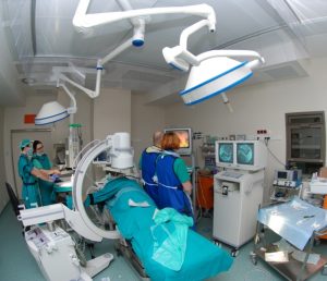 cztery osoby w ubraniach koloru niebieskiego wykonujące zabieg na bloku operacyjnym obserwują przebieg operacji na trzech monitorach koloru szarego