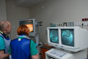 dwie osoby w ubranich koloru niebieskiego podczas operacji ogląda wykonywaną operację na trzech monitorach aparatu endoskopowego koloru białego