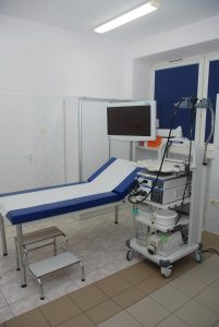 w pokoju badań endoskopowych znajduje się łóżko medyczne koloru szarego, stołek obrotowy koloru niebieskiego, na scianie ekran koloru czarnego wraz z aparaturą do badań endoskopowych