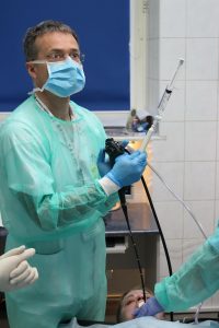 lekarz ubrany w niebieskie ubranie ochronne oraz niebieską meseczką ochronna na twarzy wykonujący badaniie gastroskopii wraz z pielęgniarka pacjentowi szpitala
