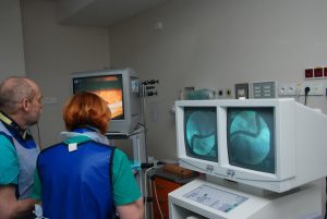 dwie osoby w ubranich koloru niebieskiego podczas operacji ogląda wykonywaną operację na trzech monitorach aparatu endoskopowego
