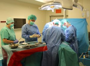 trzech lekarzy ubranych w ubrania ochronne oraz maseczki ochronne wykojuja zabieg chirurgiczny na bloku operacyjnym w asyscie dwóch instrumentariuszek przygotowujacych narzedzia do operacji na zielonym stole