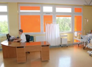 Sala chorych. W oknie pomarańczowe rolety. Pielęgniarka wprowadza dane pacjentów do komputera.