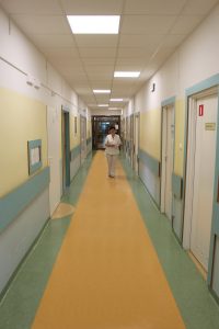 korytatarz oddziału szpitala koloru zółto zielonego i białym drzwiami idzie osoba ubrana w odzież białą ochronną