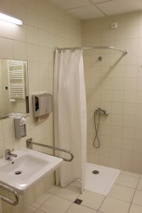 łazienka koloru białego w której znajduję się umywalka z baterią koloru białego wraz z podajnikiem mydła i papieru w kolorze srebnym z prysznicem z białym parawanem