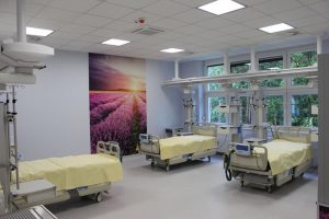 na sali oddziału OAIT koloru fioletowego znajduję się na ścianie fototapeta koloru fioletowego z zachodem słońca oraz trzy łóżka medyczne koloru biało żółtego wraz z aparaturą do ratowania życia ludzkiego