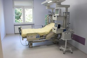 w pokoju koloru filetowego znajduję się łóżko medyczne koloru biało żółtego wyposażonę w aparaturę koloru białego do ratowania ludzkiego życia