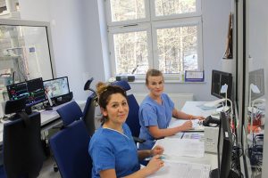 w pokoju koloru niebieskiego dwie osoby ubrane w ubrania medyczne koloru niebieskiego siedząc na fotelach obrotowych niebieskich przy biurkach z czarnymi monitorami uśmiechaja sie na oddziale OAIT
