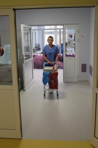 osoba stojąca w drzwiach koloru białego ubrana w ubranie ochronne medyczne koloru niebieskiego trzyma w rękach wózek medyczny koloru białego na kółkach na oddziałe OAiT