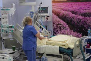 sala oddziału OAIT pielęgniarka ubrana w niebieska odzież ochronna przy pacjęcie leżącym na łóżku medycznym podpiętym do aparatury wspomagającej oddychanie obok widoczna na ścianie fototapeta koloru fioletowego