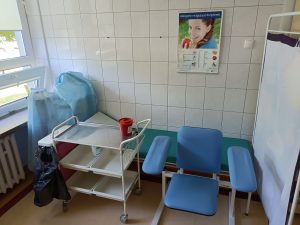 w pomieszczeniu znajduje się fotel obrotowy niebieski wraz z zieloną leżanka medyczną i białem parawanem i wózkiem medycznym na którym znajduje się czerowny pojemik medyczny.