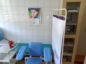 w pomieszczeniu znajduje się fotel obrotowy niebieski wraz z zieloną leżanka medyczną i białem parawanem i wózkiem medycznym na którym znajduje się czerowny pojemik medyczny.
