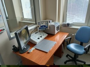 fotel obrotowy koloru niebieskiego wraz z brazowym biurkiem na którym znajduję się kokpit sterowniczy wraz z monitorem do badań RTG.