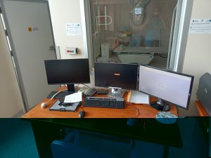 pokój techników RTG z brązowym biurkiem i sprzetem komputerowym oraz szybą podglądową do aparatu badań RTG