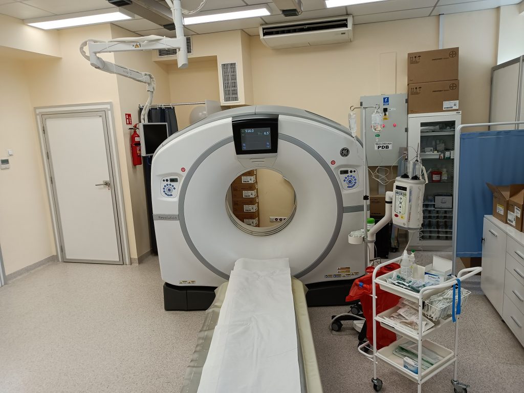 Pracownia TK wyposażona w Tomograf Komputerowy koloru białego obok wózek medyczy koloru białego wraz z prawanem koloru niebiekiego oraz drzwiami koloru białego