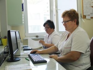dwie pielęgniarki ubrane w białe ubrania ochronne podczas pracy w systemie szpitalnym przy monitorach i klawiaturach koloru czarnego.