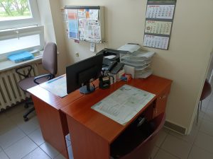 pomieszczenie poradni chirurgicznej wyposażoen w dwa biurka koloru brązowego na którym znajduję się sprzęt komputerowy obok fotel obrotowy koloru brązowego