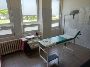 pomieszczenie koloru białego w którym znajduję się leżak medyczny koloru niebieksiego wraz z wózkiem medycznym koloru białego