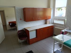 pomieszczenie poradni chirurgicznej wyposażone meblosciankę koloru brązowego oraz dwa krzesełka koloru brązowego oraz leżakmedyczny koloru niebieskiego.