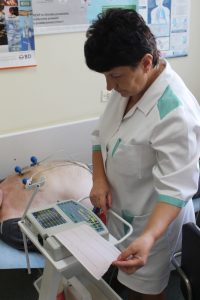 osoba ubrana w medyczne ubranie ochronne koloru białego wykonuje badania ekg na aparacie EKG