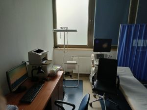 Pomieszczenie pracowni USG wyposażone w fotel obrotowy koloru czarnego wraz z leżakiem medycznym kolory niebieskiegi oraz aprataem do badań USG oraz biurkiem ze sprzetem komputerowym i drukarką.