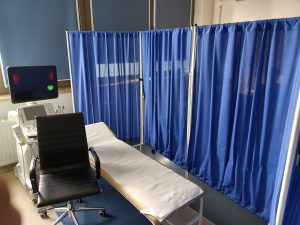 Aparat do badań USG obok którego znajduję się łózko medyczne koloru białem z parawanami koloru niebieskiego i fotelem obrotowym koloru czarnego.