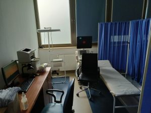 Pomieszczenie pracowni USG w którym znajduję się łóżko medyczne koloru białegoz niebieskim parawanem wraz z aparatem do badań usg, fotelem obrotowym kolory czarnego i biurkiem na którym znajduje sie sprzęt komputerowy z drukarką.