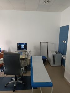 Pomieszczenie pracowni USG wyposażone w fotel obrotowy koloru czarnego wraz z leżakiem medycznym kolory niebieskiegi oraz aprataem do badań USG.