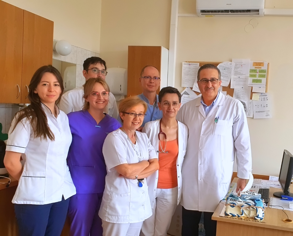 Personel lekarski. Cztery kobiety oraz trzech mężczyzn, wszyscy uśmiechnięci pozują do zdjęcia. Jedna posiada stetoskop na szyi. Przebywają w pokoju lekarskim.
