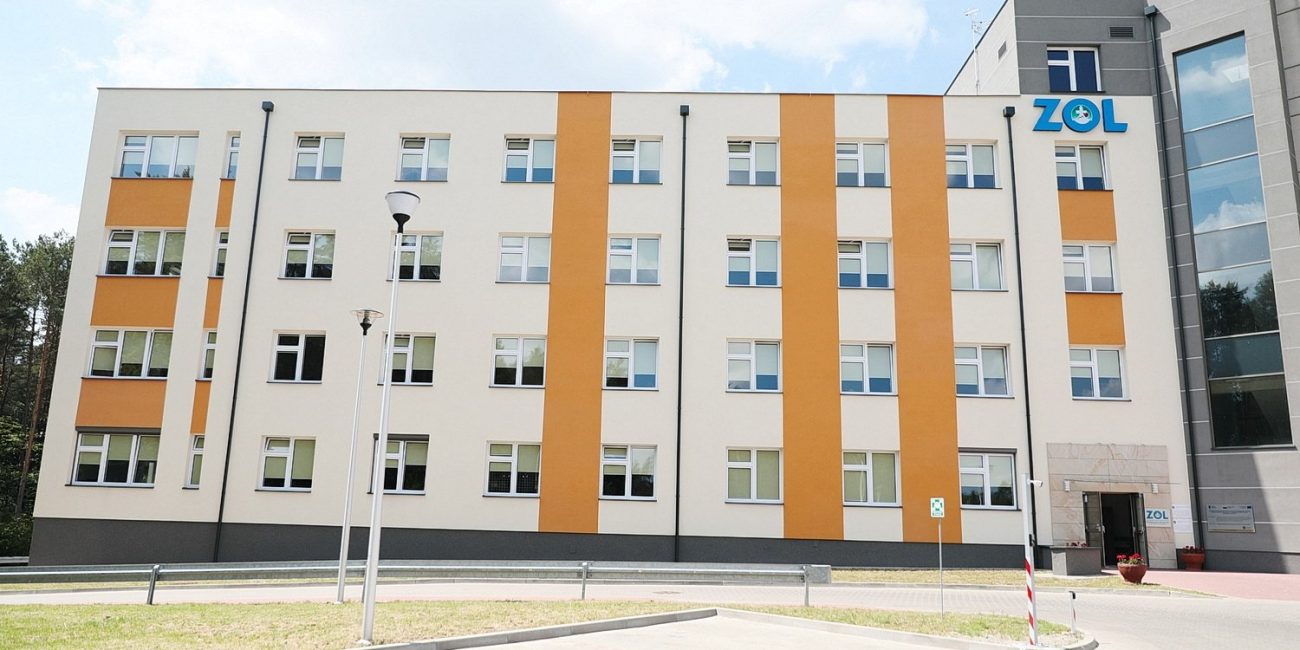 Budynek ZOL koloru białego z pomaranczowymi pasami, w górnym rogu po prawej stronie znaduję sie niebieskie logo ZOL.