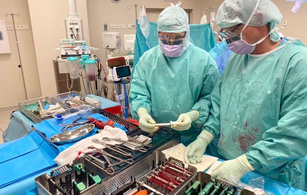 Fotografia przedstawia dwóch chirurgów podczas operacji. Na zdjęciu widzimy również salę i medyczne wyposażenie
