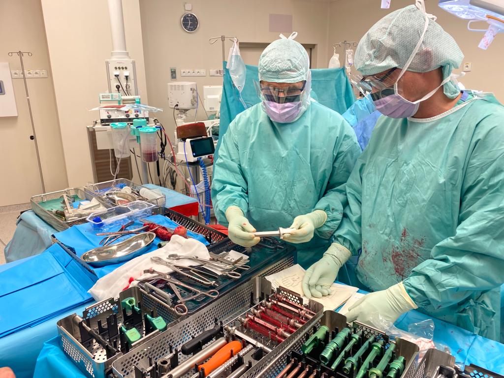 Fotografia przedstawia dwóch chirurgów podczas operacji. Na zdjęciu widzimy również salę i medyczne wyposażenie