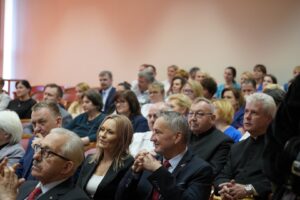 Fotografia przedstawia grupę osób siedzących i słuchających przemówienia