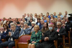 Fotografia przedstawia grupę osób siedzących i słuchających przemówienia z biskupem na pierwszym planie